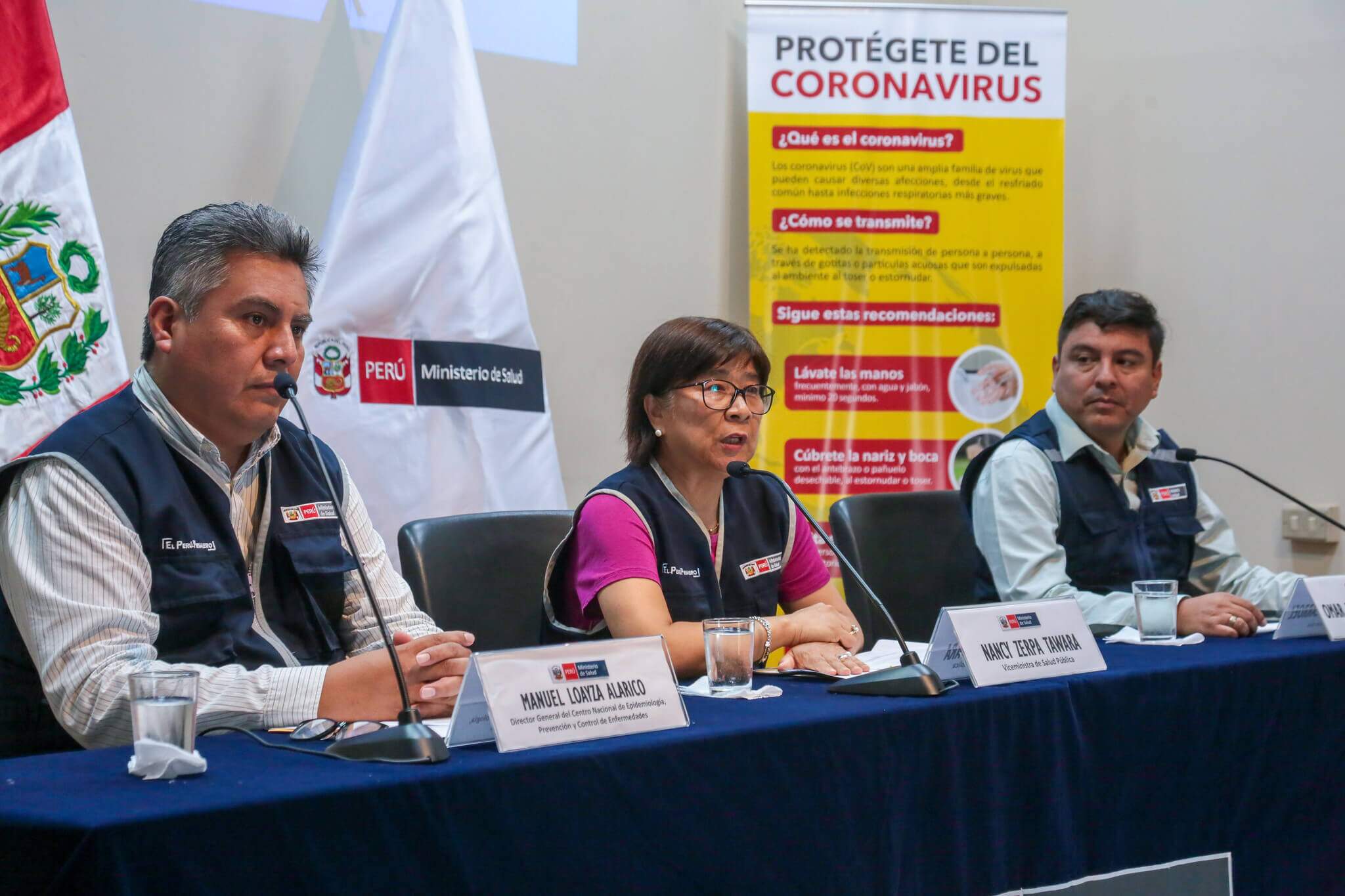Três agentes do ministério da saúde do Peru sentados apresentando o debate sobre o Coronavírus. Ao fundo, uma placa com as informações do vírus em espanhol. Conteúdo sobre Estado de Emergência.