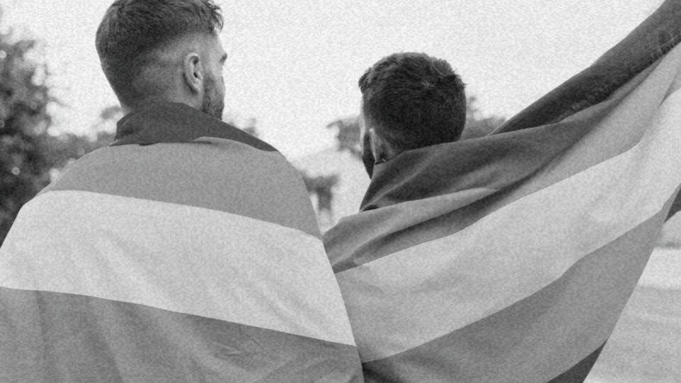 Imagem de um casal homossexual de costas enrolados em bandeiras LGBTQIAP+ representando os direitos LGBT+