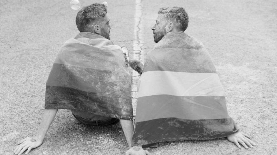Imagem de duas pessoas homossexuais sentados com a bandeira da comunidade LGBTQIAP+ representando a LGBTfobia e o desafios dessa população