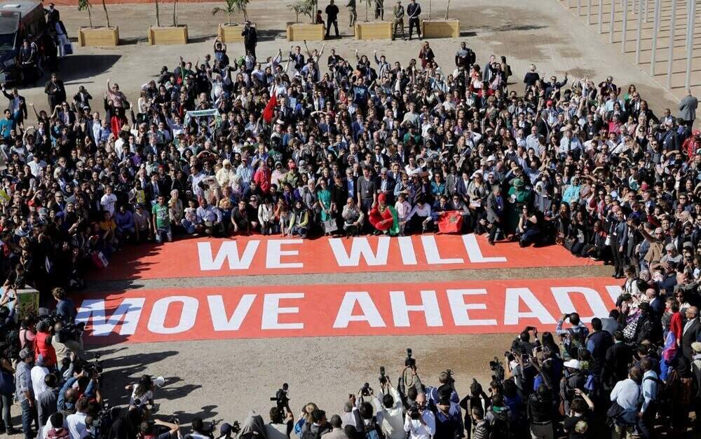Pessoas estendem faixa com os dizeres "we will move ahead" no chão durante a COP 22. Imagem do conteúdo sobre Aquecimento global.