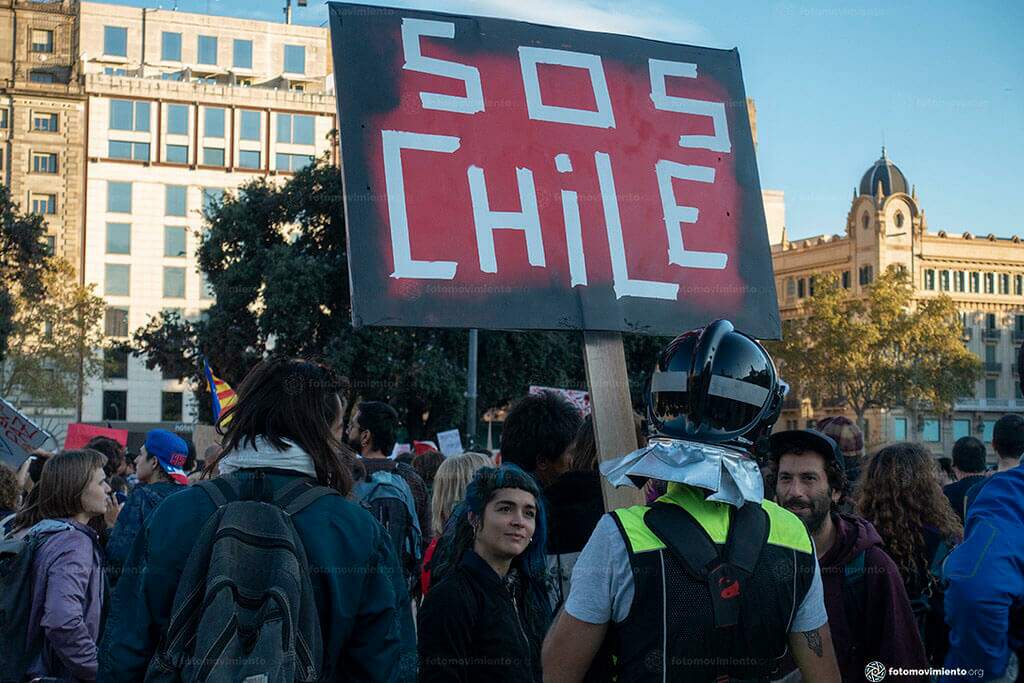 Manifestantes na rua com placa SOS CHILE. Conteúdo sobre crise econômica do Chile.