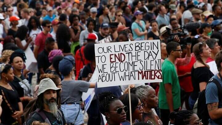 Na imagem, manifestantes e uma mulher segurando uma placa "our lives begin to end the day we becomes silent about what matters". Conteúdo sobre Discurso de ódio.