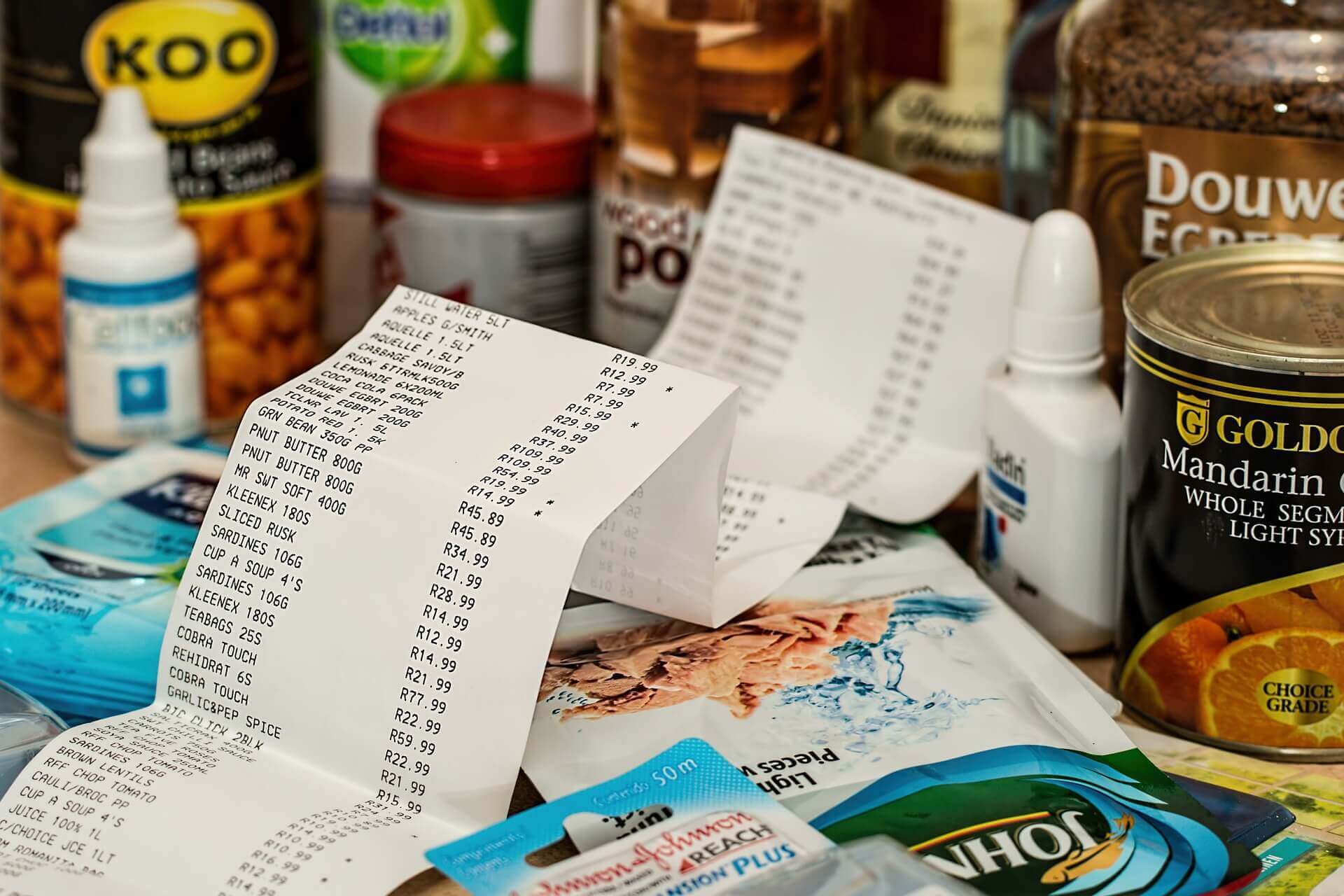 No centro da imagem, uma nota fiscal de uma grande compra. Em volta do papel, vários produtos de alimentação e higiene pessoal