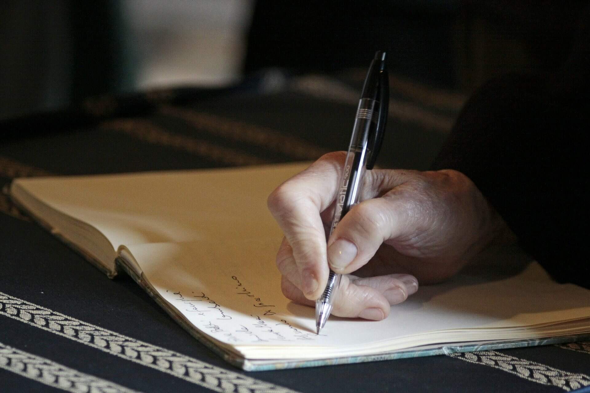 Mão direta de uma pessoa com uma caneta transparente, de tinta preta, escrevendo em uma agenda sem linhas sob uma mesa com pano preto com detalhes em branco | Direito de herança – Artigo Quinto