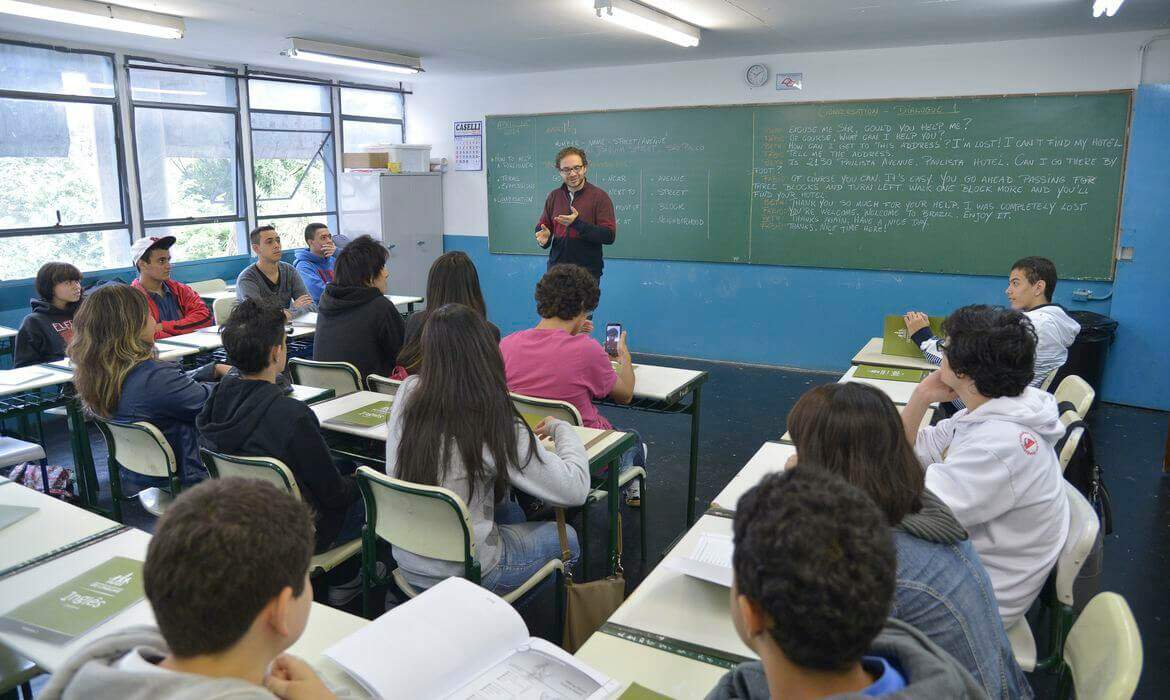 Na imagem, professor dando aula para diversos alunos em sala de aula.