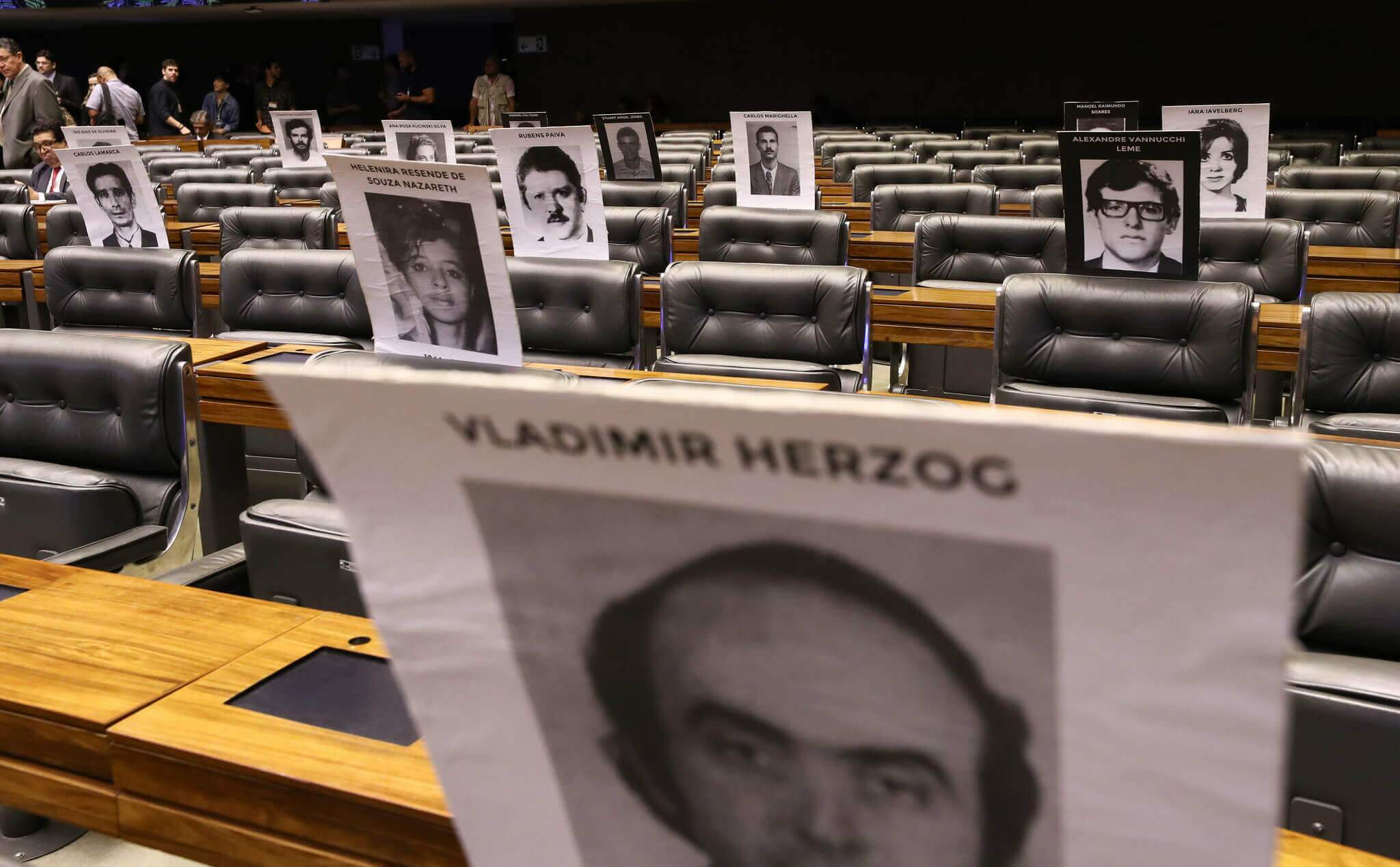 Cadeiras do Congresso Nacional vazias com imagens impressas em memória daqueles perseguidos durante a ditadura militar. Conteúdo sobre AI-5