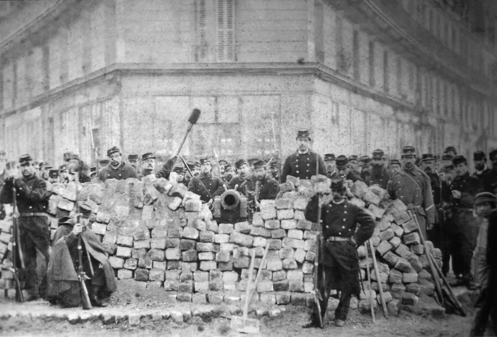 A Barricada em Paris, no ano de 1871. Vários militares posicionados atrás de um canhão e um pequeno muro de pedras. Texto: O que foi a Comuna de Paris?