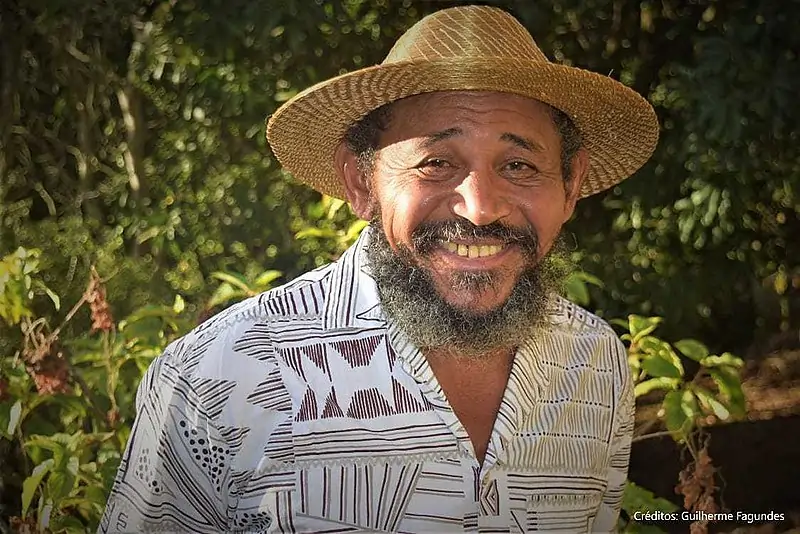 Foto de Nêgo Bispo, pensador quilombola e precursor do contracolonialismo, sorrindo, com blusa estampada e chapéu de palha, à frente de arbustos verdes.