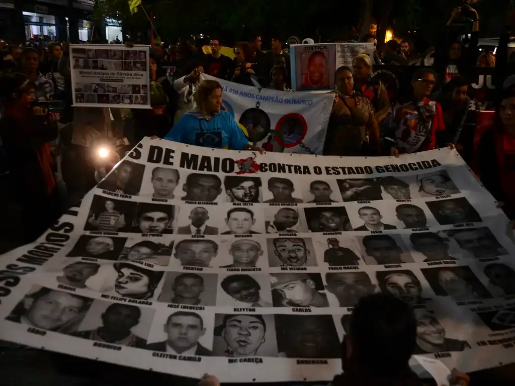 A imagem mostra uma das Mães de Maio carregando um cartaz com fotos das vítimas dos Crimes de Maio estampadas, durante uma manifestação