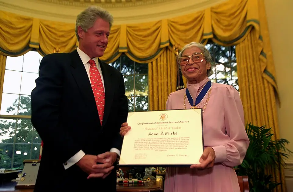 foto colorida de um homem branco, cabelos grisalhos e terno entregando um certificado a uma mulher negra de cabelos grisalhos amarrado para trás, de ocúlos e vestido rosa.