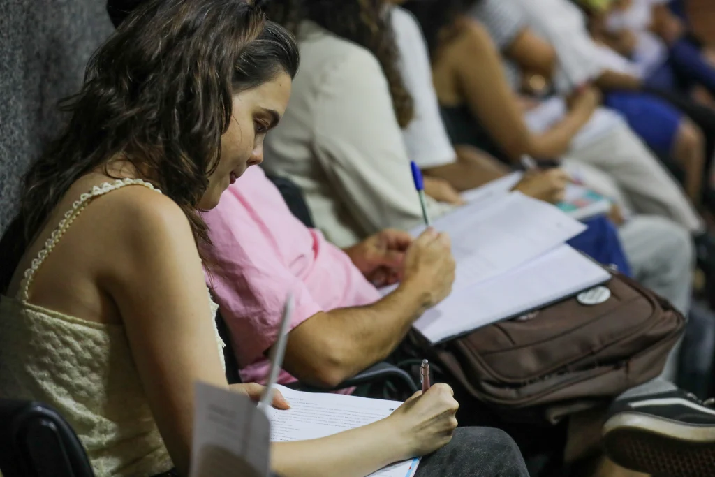 Mulher jovem sentada, preenchendo um documento, em uma sala cheia de pessoas ao fundo, semelhante ao que acontece durante uma prova de concurso público.