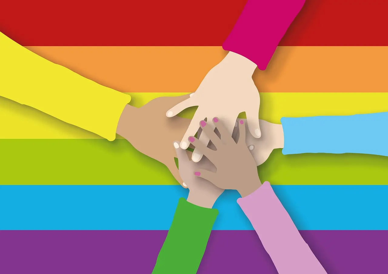Representação gráfica de mãos unidas, ao fundo, as cores da bandeira LGBTQIA++. Texto: O que é o nome social e como surgiu?