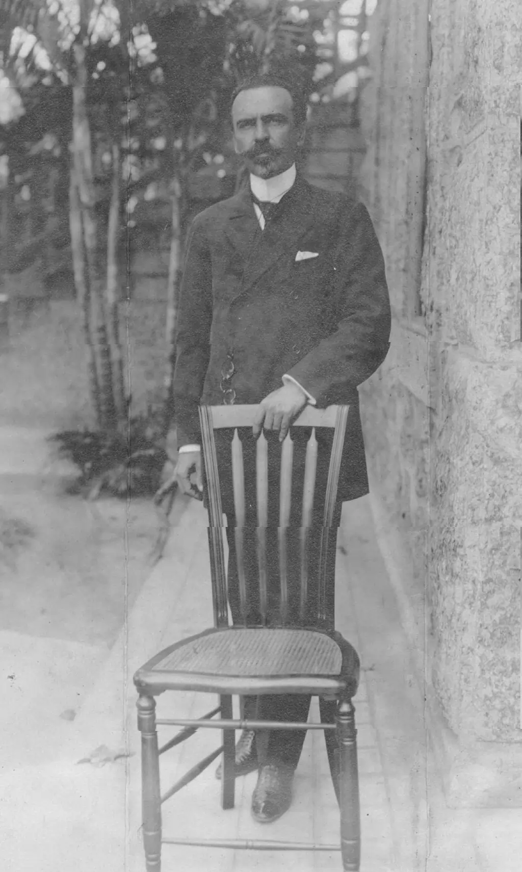 Nilo Peçanha segurando a cabeceira de uma cadeira de madeira, em pé.