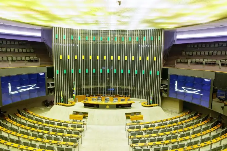 Vista interna do plenário da Câmara dos Deputados, em Brasília, com cadeiras dispostas em semicírculo, a mesa diretora ao centro e o painel de madeira com luzes verdes e amarelas ao fundo.