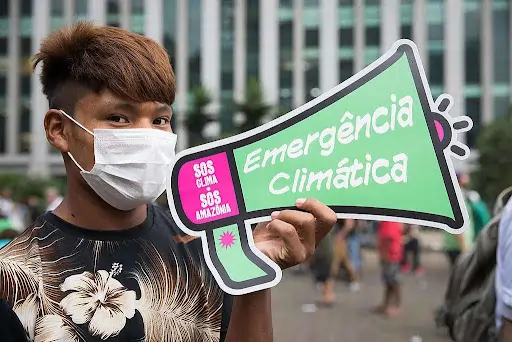 A foto mostra uma pessoa usando máscara de proteção enquanto segura um cartaz colorido em formato de megafone, com as frases "SOS Clima", "SOS Amazônia" e "Emergência Climática". Ao fundo, há pessoas e o que parece ser a estrutura de um prédito alto. Texto: Você sabe o que é a Política Nacional de Educação Ambiental (PNEA)?