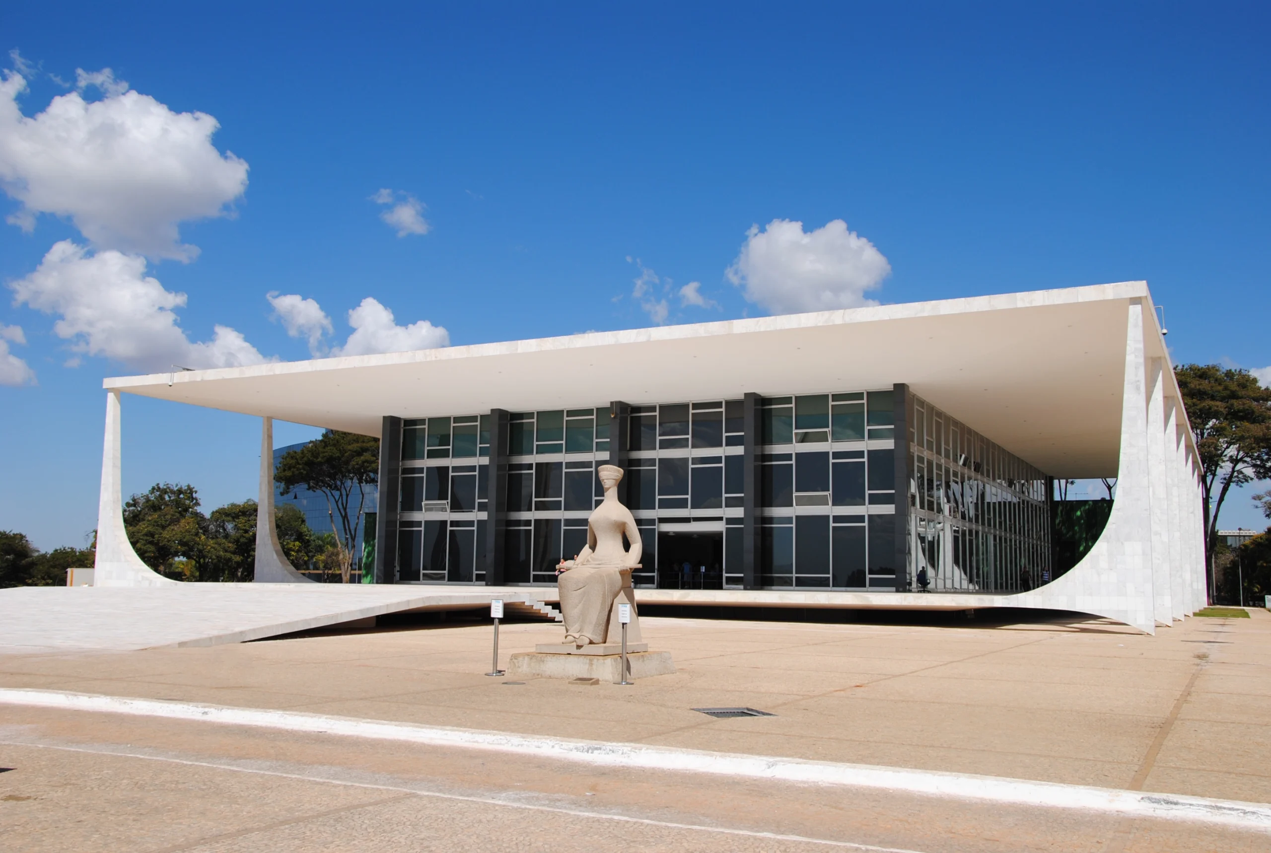 Foto do prédio do STF, localizado na Praça dos Três Poderes, em Brasília. A construção, projetada por Oscar Niemeyer, tem fachada com colunas retas e, em frente, a icônica estátua da Justiça – uma figura sentada com os olhos vendados. Texto: Ministros do Brasil: o caminho até os Tribunais Superiores