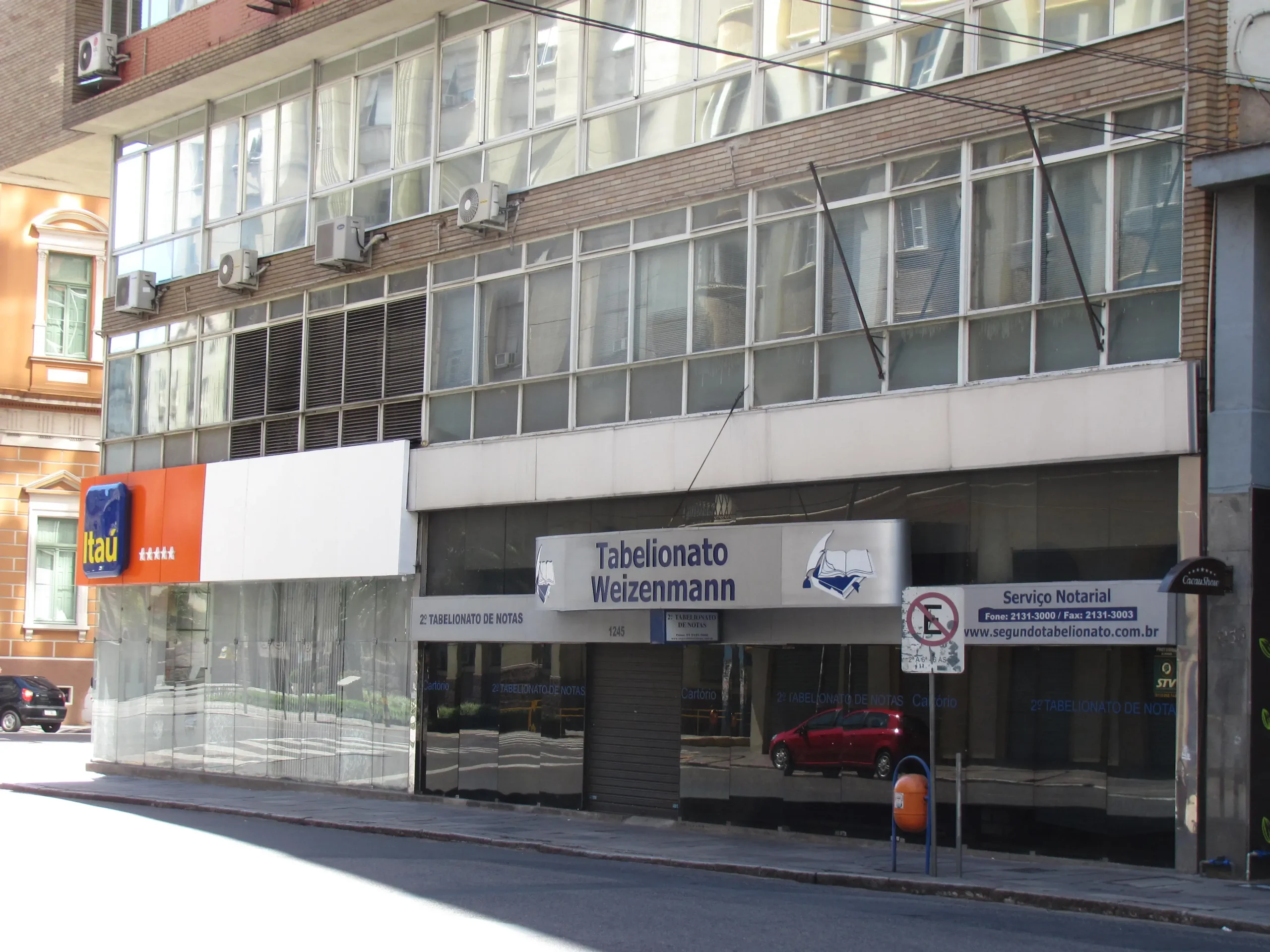 Fachada com a placa ‘Tabelionato Weizenmann’ e identificação de serviços notariais, localizada em prédio comercial ao lado de uma agência do banco Itaú.
