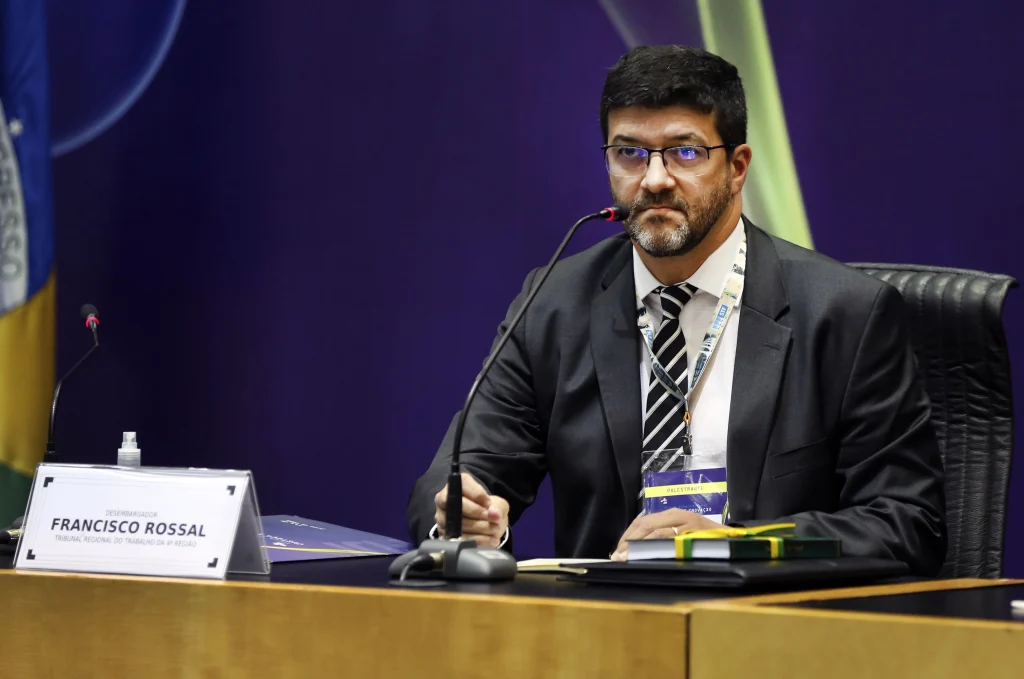 Homem de terno, usando óculos, sentado à mesa com crachá no pescoço e microfone à frente, durante um evento oficial no Plenário do Tribunal Superior do Trabalho. 