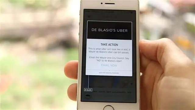 Uma mão segurando um smartphone branco que exibe uma tela do aplicativo Uber com o modo “De Blasio’s Uber”. No centro da tela há uma caixa de aviso com o título “TAKE ACTION”, explicando que é assim que o Uber ficaria em Nova York se o projeto de lei do prefeito De Blasio fosse aprovado, e incentivando o usuário a enviar um e-mail para o prefeito e o conselho municipal dizendo “NÃO” ao projeto.