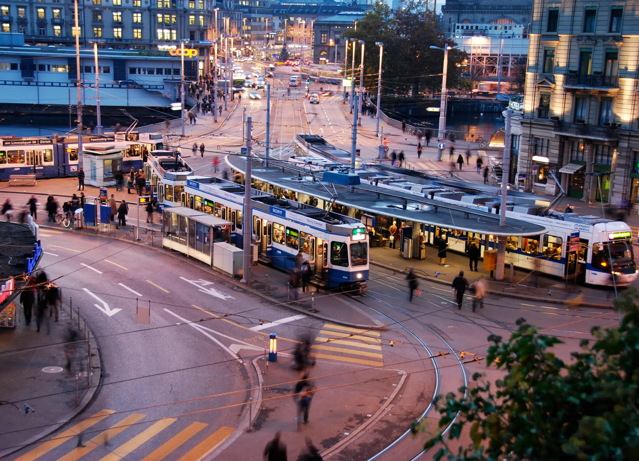 Vista aérea de uma movimentada praça de transporte público em Zurique ao entardecer. Vários bondes azuis e brancos chegam e partem de plataformas cheias de pessoas. As ruas curvam-se ao redor dos trilhos, com pedestres caminhando rapidamente. Prédios iluminados, lojas e luzes urbanas compõem o cenário, transmitindo a atmosfera organizada e dinâmica da cidade.