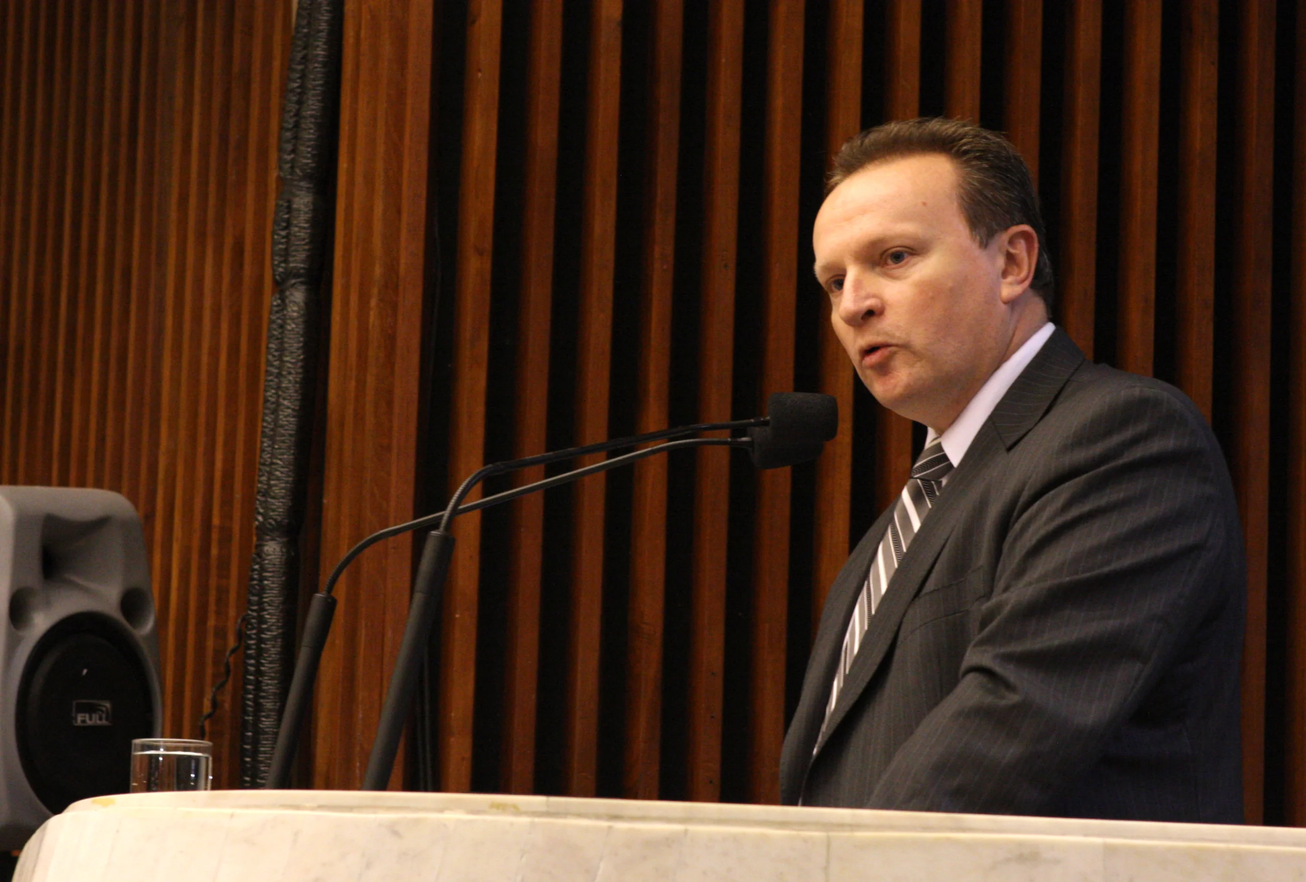 Homem branco, de pé, vestindo terno preto e gravata listrada em preto e branco, fazendo um pronunciamento ao microfone.