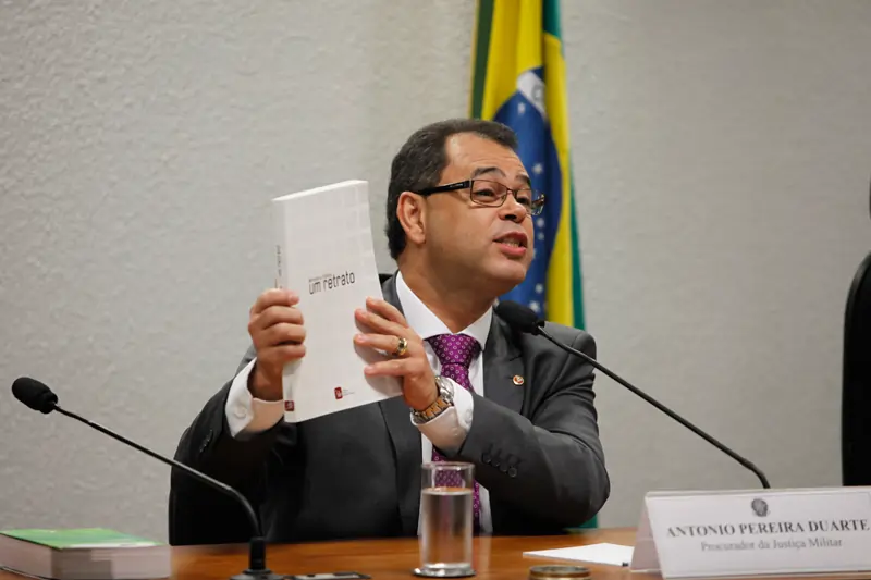 Homem de óculos e terno cinza, identificado por uma placa como Antônio Pereira Duarte, Procurador da Justiça Militar, segura um livro branco com o título "Ministério Público: um retrato". Ele está sentado à mesa com microfones e a bandeira do Brasil ao fundo.