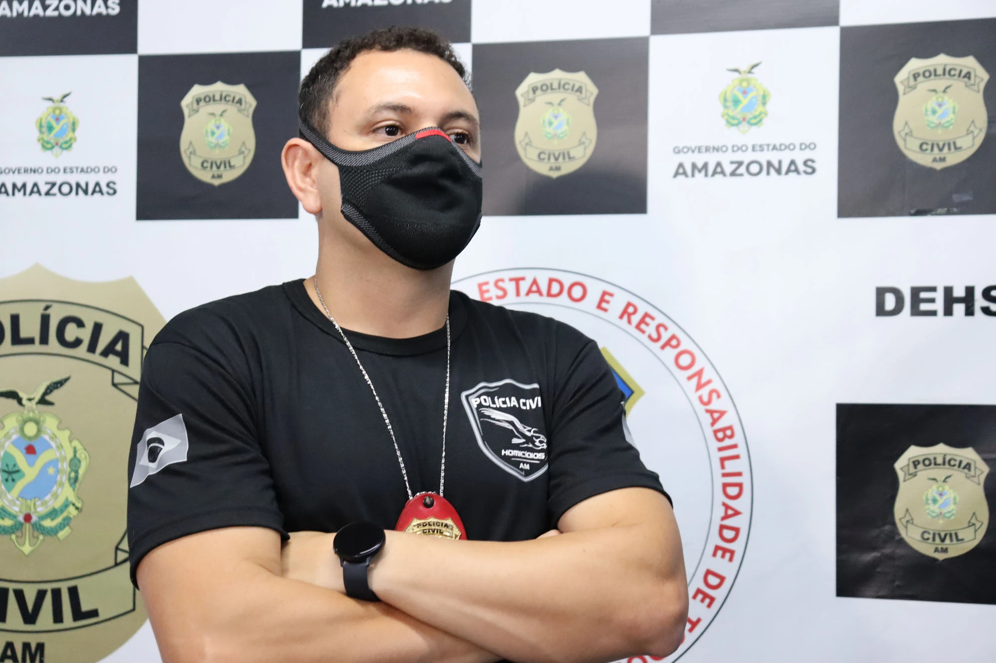 Homem usando máscara preta de proteção facial, camiseta preta com o emblema da Polícia Civil - Homicídios/AM e distintivo vermelho pendurado no pescoço, de braços cruzados. Ao fundo, há um painel com logotipos da Polícia Civil e a inscrição "Governo do Estado do Amazonas".