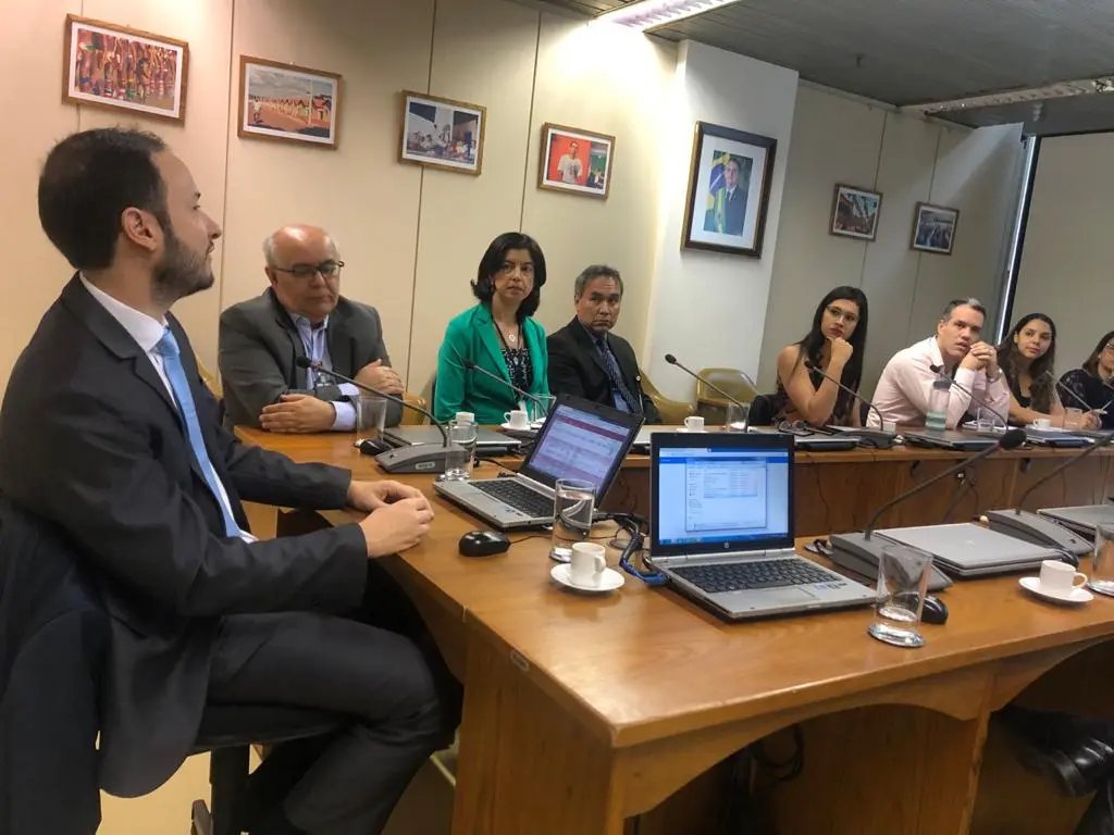 Grupo de nove pessoas, entre homens e mulheres, utilizando roupas formais de trabalho, sentados em uma mesa de conferência, com dois computadores portáteis abertos sobre a mesa.